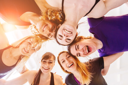 Team of young laughing group of teenager fitness girls hugging together after workout on white background. Underneath view, sun glare effect.