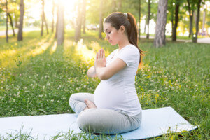 Young healthy pregnant woman doing yoga exercises in nature outdoors on green grass on fitness mat at sunset. Happy pregnancy and motherhood concept