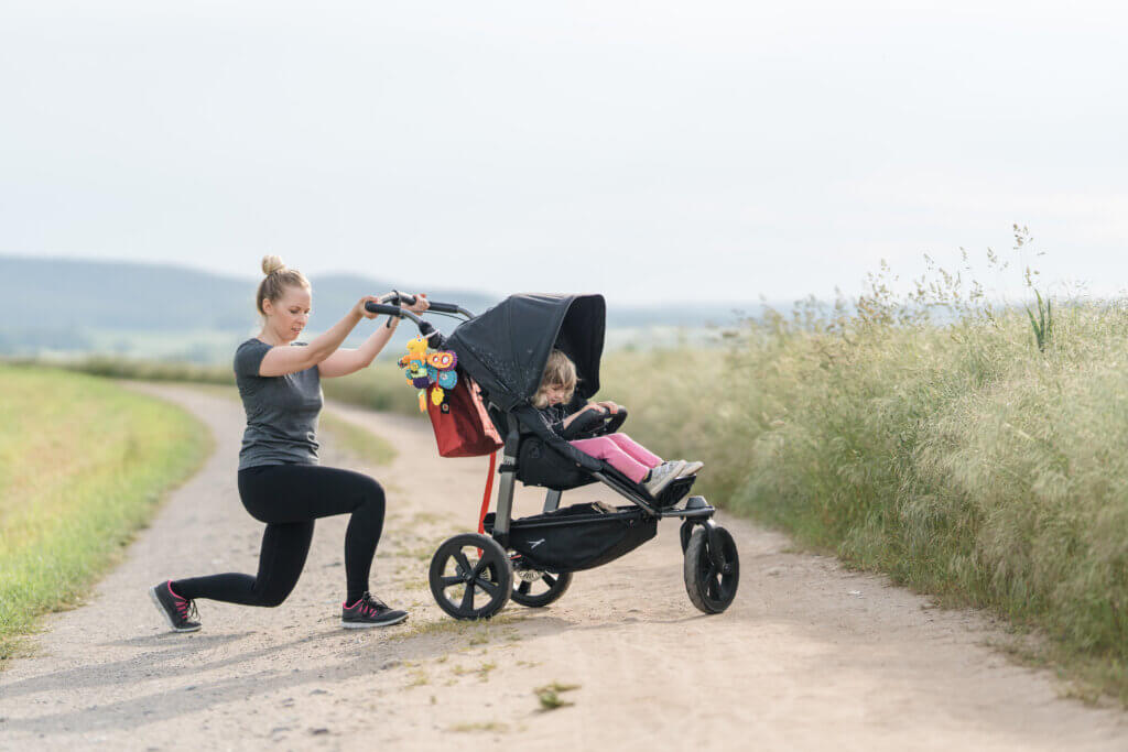 Sportliche Mama am Kinderagen, macht einen Ausfallschritt. Kind sitzt im Buggy,