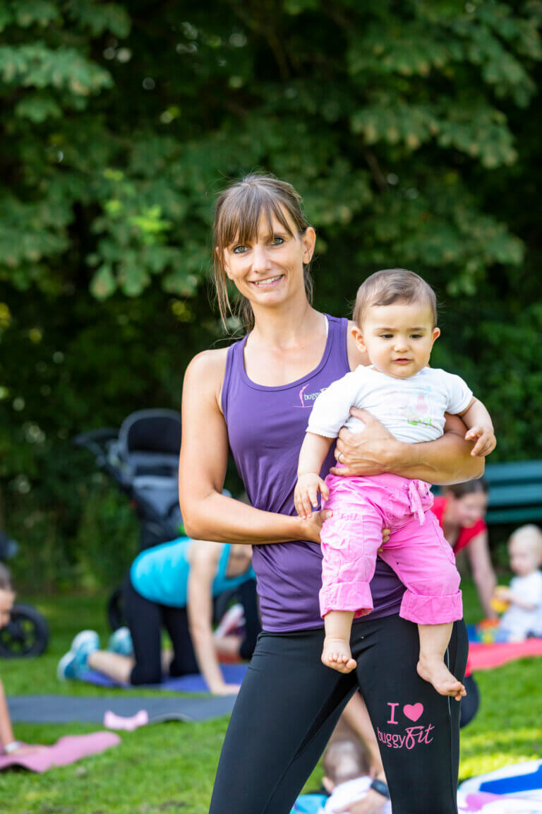 buggyFit Trainerin mit Baby auf dem Arm. Im Hintergrund trainieren Mamas.