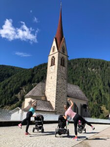 2 Mamas machen Sport mit Kinderwagen vor einer Kirche.