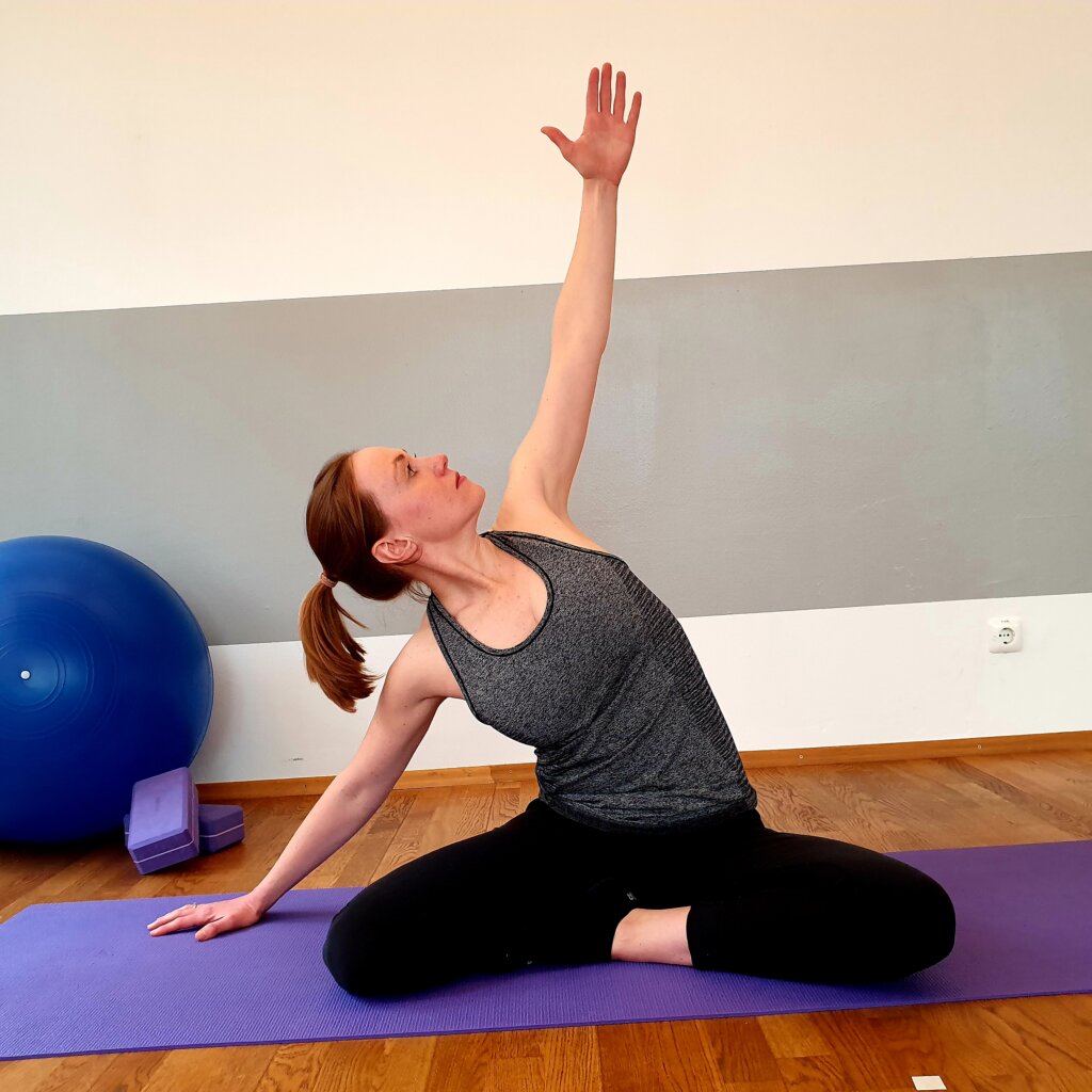 Frau macht eine Pilates-Uebung im Sitzen auf der Matte, daneben Gymnastikball und Bloecke