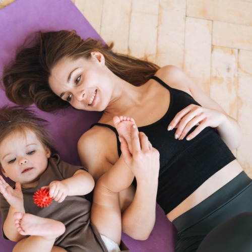 Mama liegt mit ihrem Baby auf einer Yogamatte, das Baby spielt mit einem roten Igelball