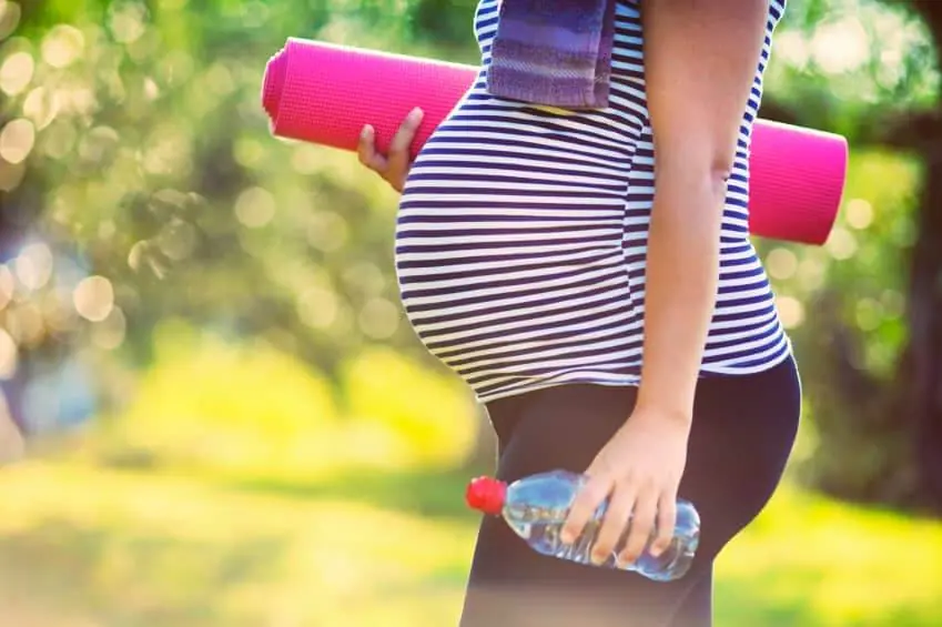 Eine Schwangere mit Babybauch geht mit Yogamatte und Wasserflasche im Park spazieren