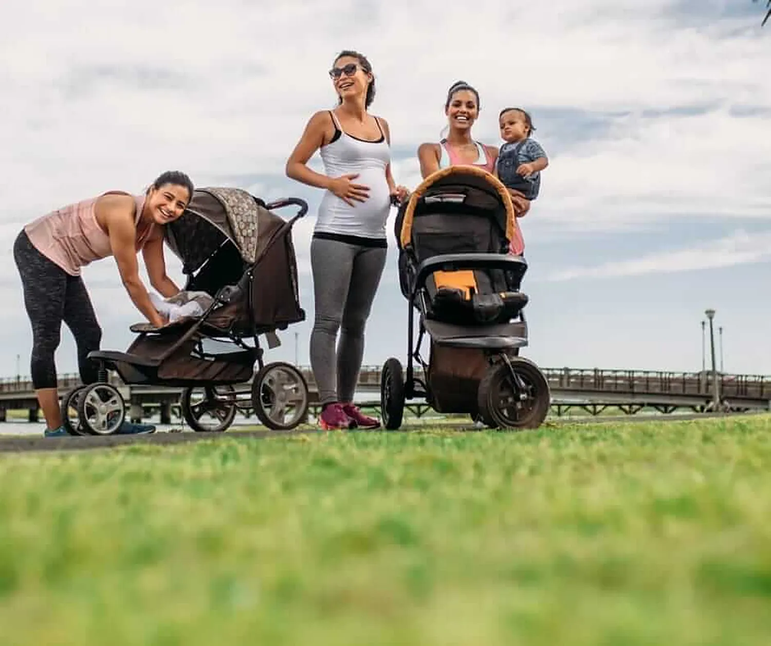 Eine Schwangere und zwei Mamas mit Kinderwagen und Baby stehen lachend im Park zusammen