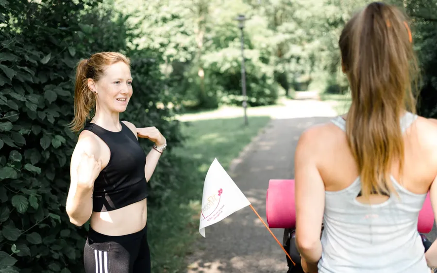 Eine buggyFit-Trainerin zeigt im Park eine Übung, vor ihr steht eine Teilnehmerin mit Kinderwagen und Matte, daneben die buggyFit-Fahne