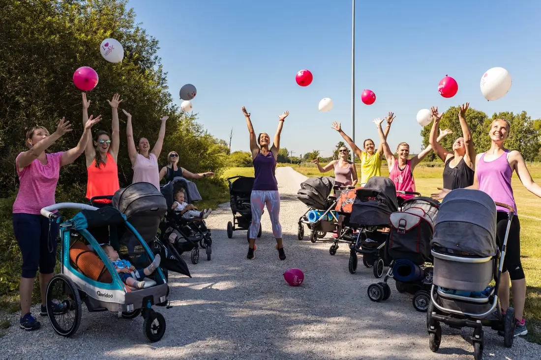 Eine Trainerin und Mamas werfen im Park pinke und weiße Luftballons in die Luft, dazwischen Kinderwagen