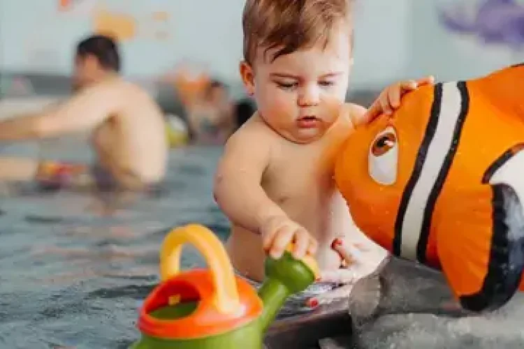 Baby spielt im Wasser in einem Babybecken mit Gießkanne und Fisch.